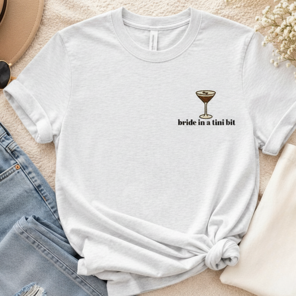 White t-shirt with 'bride in a tini bit' text and cocktail graphic on a beige surface with jeans and a hat.